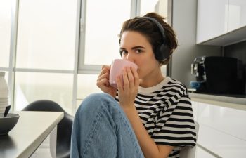 Close up portrait of smiling brunette woman, student drinks her tea and listens music or ebook in headphones, sits in kitchen and relaxes.