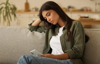 Boring sad upset frustrated stressed Arabian woman Indian girl female Hispanic lady exhausted sorrowful disappointed holding mobile phone smartphone unwanted pregnancy problem thinking at home couch